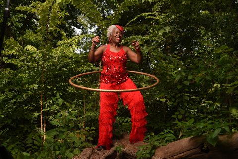 Retiree Denise Williams wearing a vibrant red outfit, smiling, and hula hooping in front of some greenery.