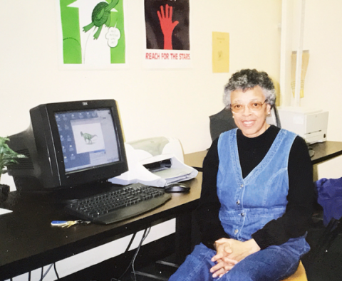Photo of Retiree Edwina R. Justice. She is in a classroom in front of a desktop computer. She is sitting down wearing a denim dress and reading glasses.